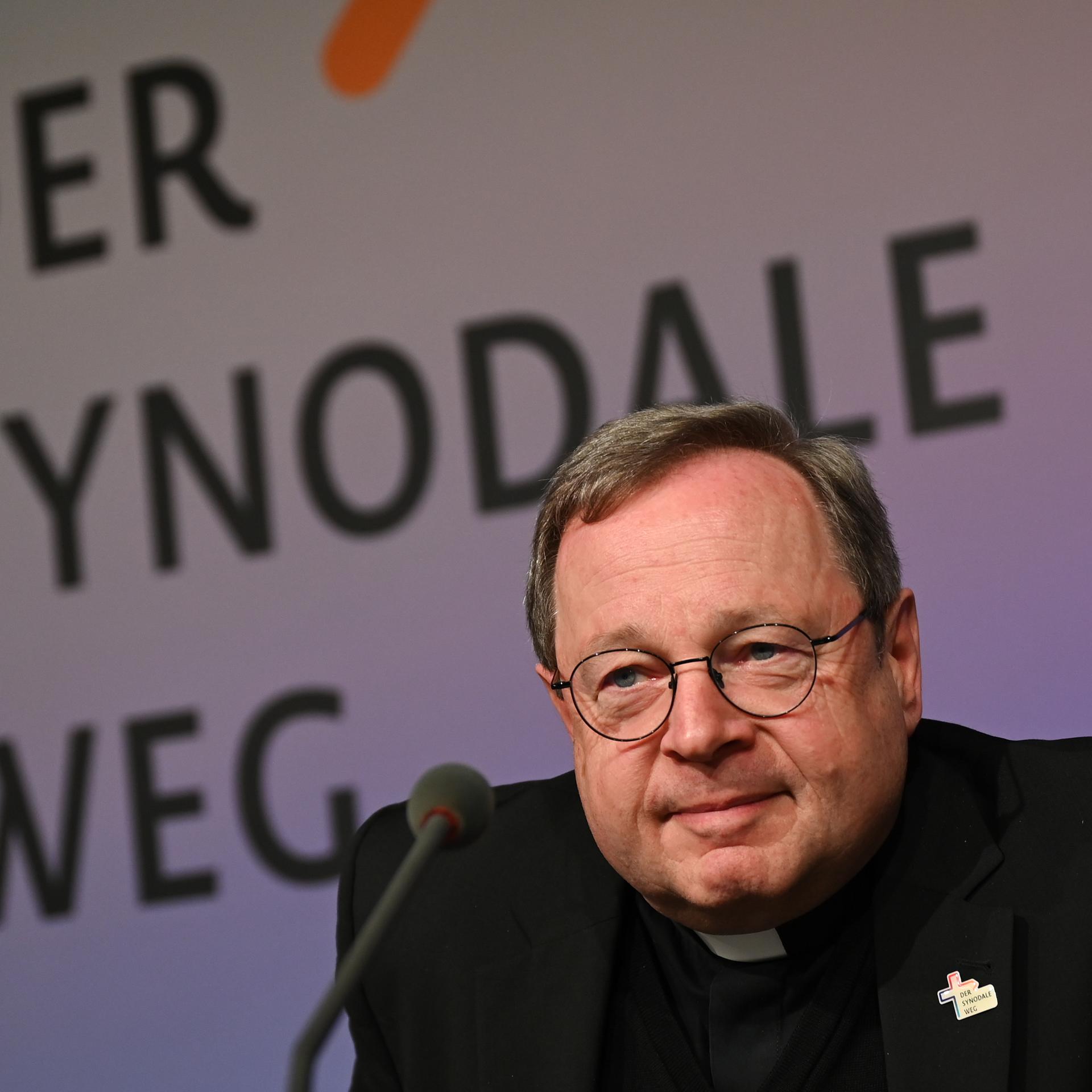 Georg Bätzing, Bischof von Limburg und Vorsitzender der Deutschen Bischofskonferenz, mit Brille und einem Kreuz am Revers, sitzt hinter einem Mikrofon und vor einem Schild, auf dem "Der synodale Weg" steht.