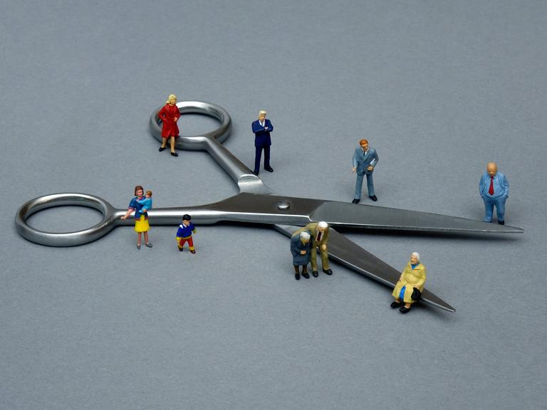 Ein Symbolbild zeigt um eine Schere gruppierte kleine Figuren, um so die Teilung der Gesellschaft zu veranschaulichen.