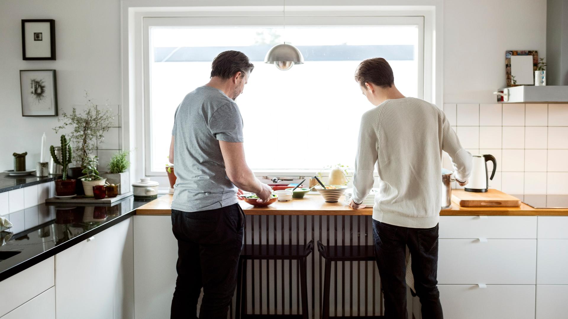 Rückansicht von Vater und älterem Sohn bei der Zubereitung von Speisen in der Küche ihres Hauses.