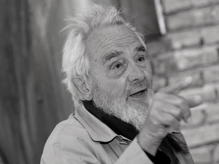 Das schwarz-weiß-Bild zeigt Arnulf Rainer. Er steht vor einer gemauerten Wand und bedeutet mit seiner rechten Hand und dem ausgestreckten Zeigefinger in eine Richtung.