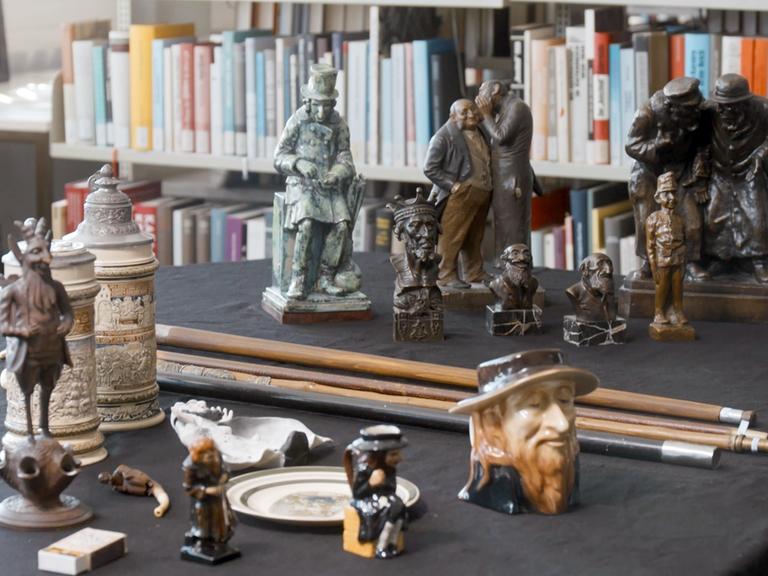 Auf einem Schreibtisch stehen verschiedene Objekte, die unter anderem menschliche Figuren in antisemitisch-klischeehafter Weise darstellen. 