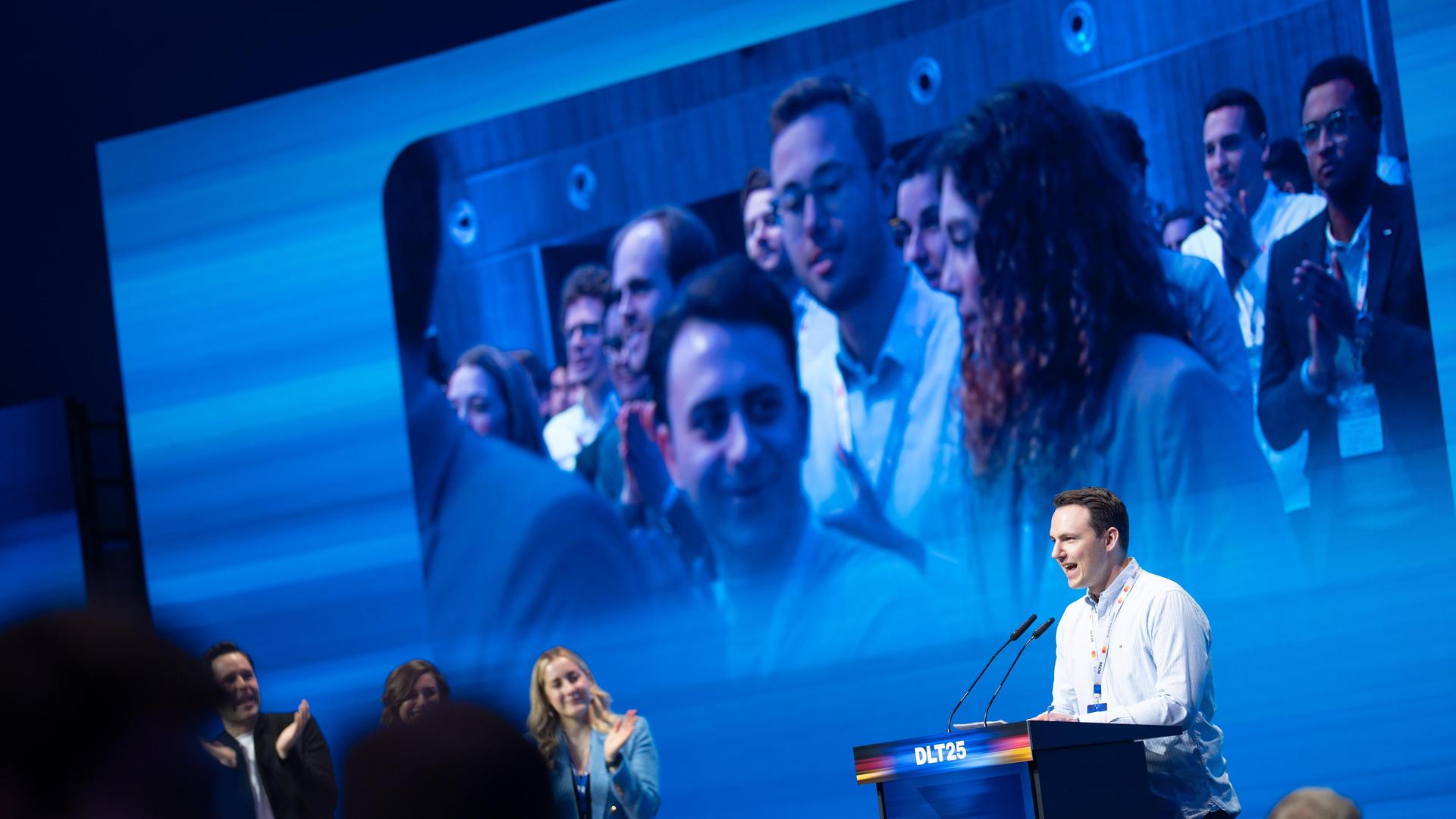 Pascal Reddig steht auf der Bühne an einem Rednerpult. Er trägt ein weißes Hemd. Im Hintergrund ist eine Leinwand mit einem Foto von mehreren JU-Mitgliedern.