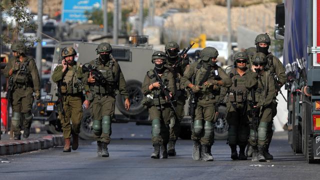 Mehrere israelische Soldaten gehen neben- und hintereinander eine Straße in der Stadt Hebron im West-Jordan-Land entlang. Dahinter ein Militärfahrzeug. 