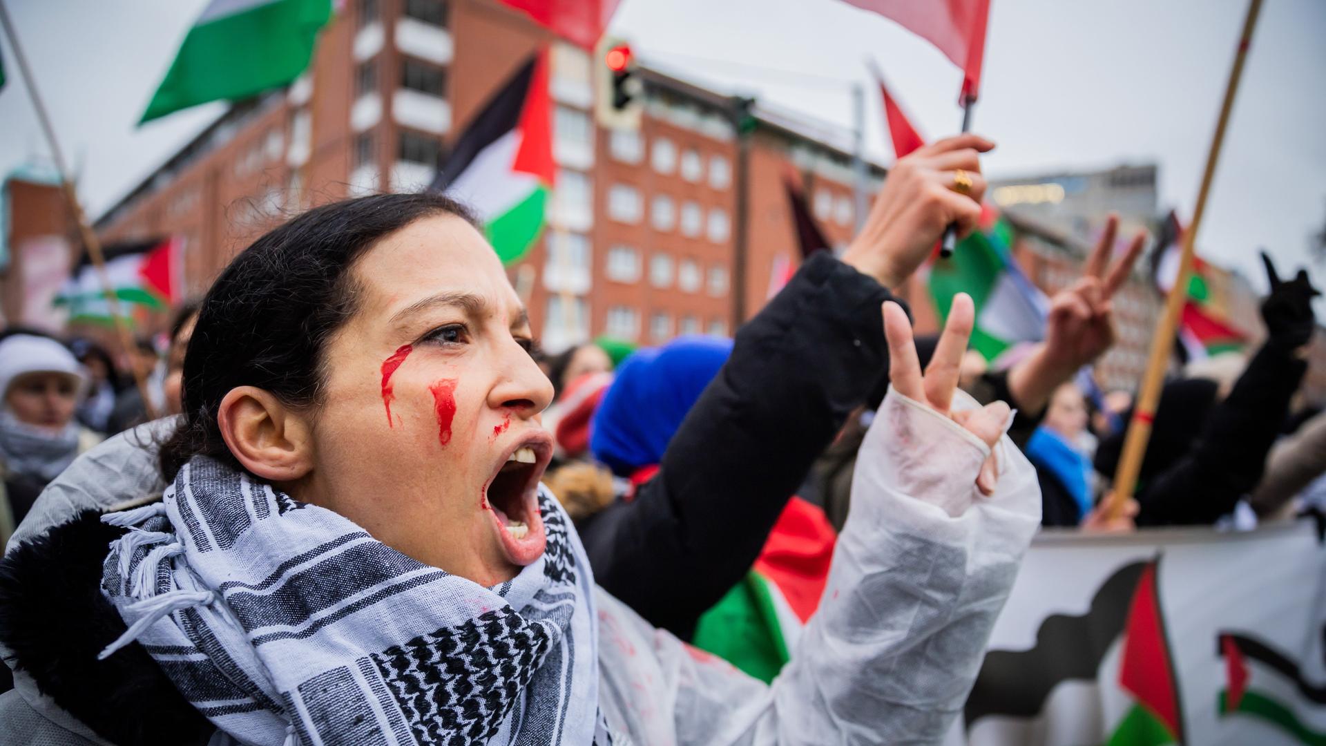 Die schwarzhaaarige Frau im Regencape trägt einen Palästinenserschal und hat sich rote Farbe ins Gesicht geschmiert. Sie macht mit der linken Hand das "Victory"-Zeichen und ruft etwas. Dahinter unscharf weitere Demonstranten und Palästina-Fahnen sowie ein Gebäude aus rotem Backstein.