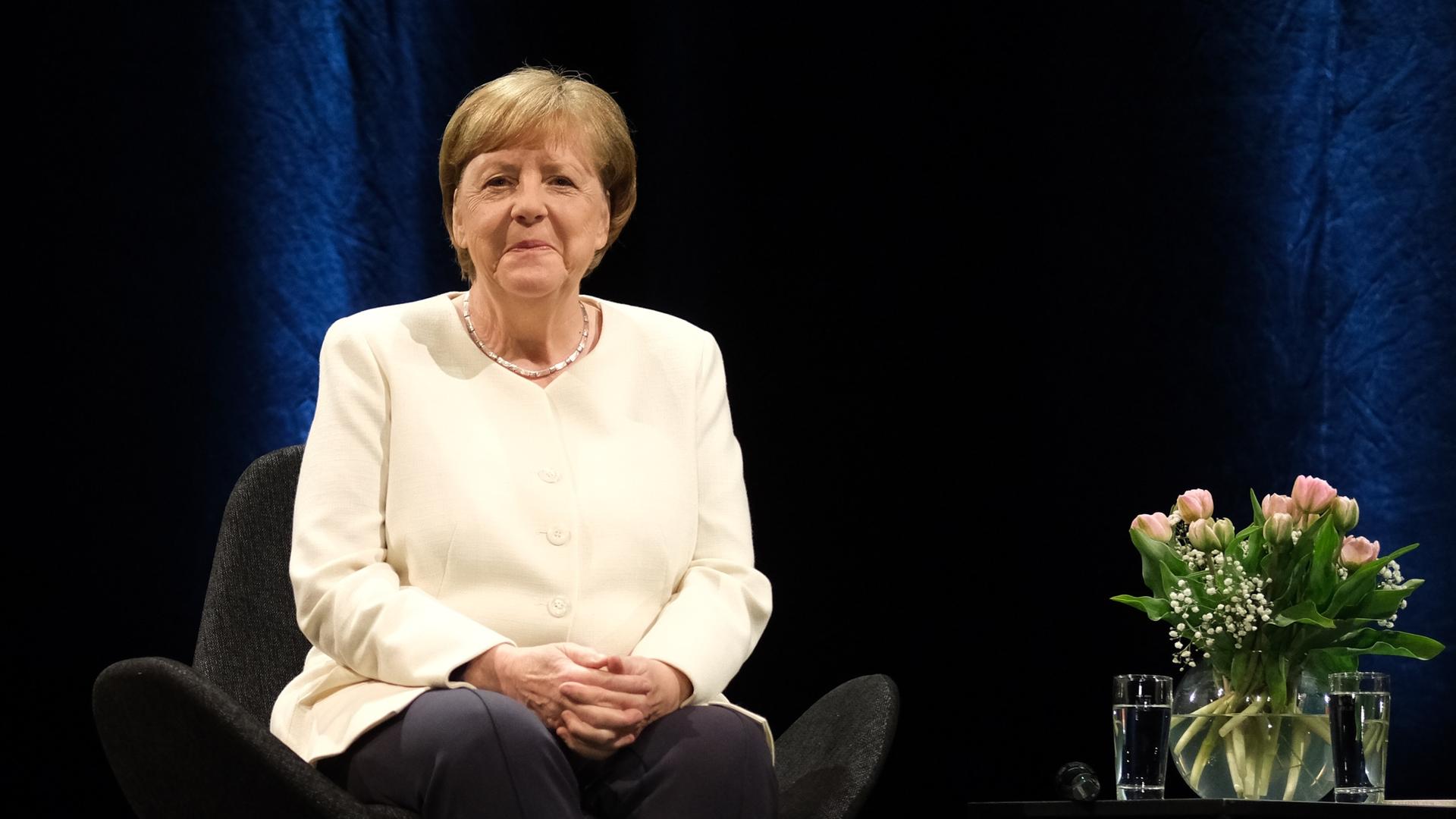 Die frühere Bundeskanzlerin Angela Merkel sitzt auf einem Stuhl vor dunklem Hintergrund, neben ihr ein Tischchen mit einem Blumenstrauß.