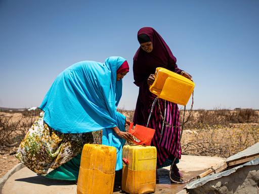 Zwei Frauen, in bunte Tücher gehüllt, füllen an einem Brunnen unter freiem Himmel Wasser in Platikkanister ab.