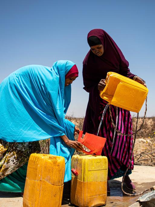 Zwei Frauen, in bunte Tücher gehüllt, füllen an einem Brunnen unter freiem Himmel Wasser in Platikkanister ab.