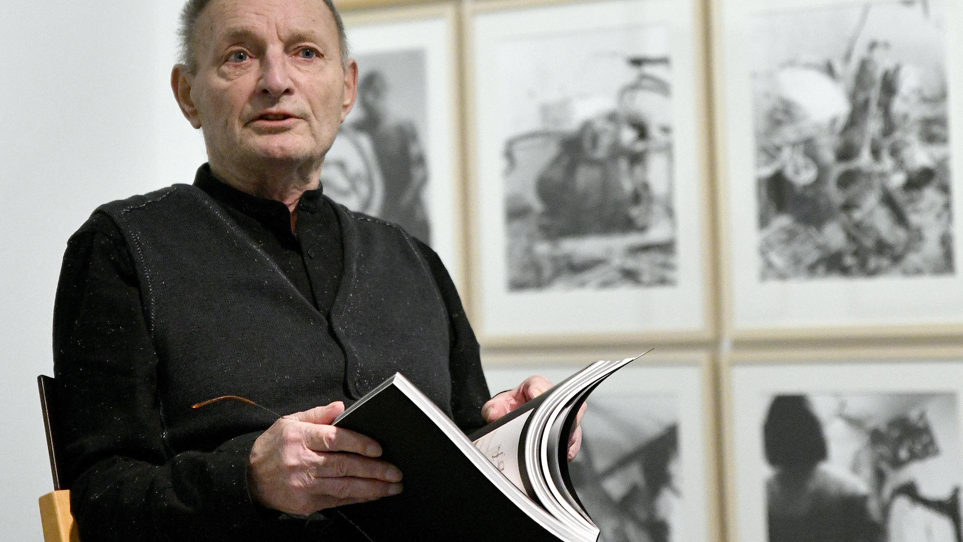 Günter Brus sitzt bei einer Presseführung zu einer Ausstellung vor seinen Bildern. Er ist ein älterer Mann mit kurzem Haar in dunkler Kleidung.