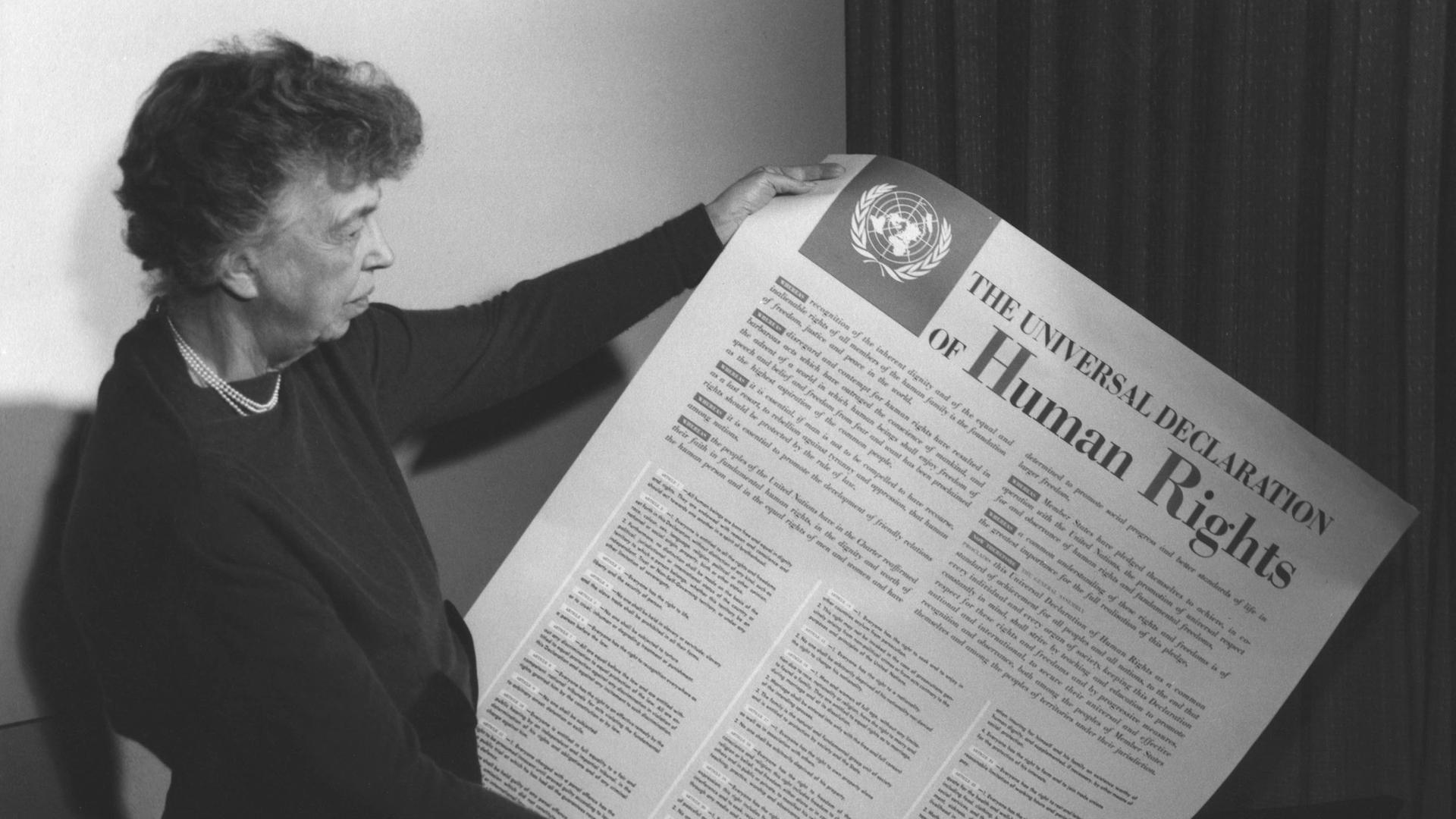 Ein Schwarz-Weiß-Foto zeigt Eleanor Roosevelt, wie sie die Allgemeine Erklärung der Menschenrechte der Vereinten Nationen hält und betrachtet.