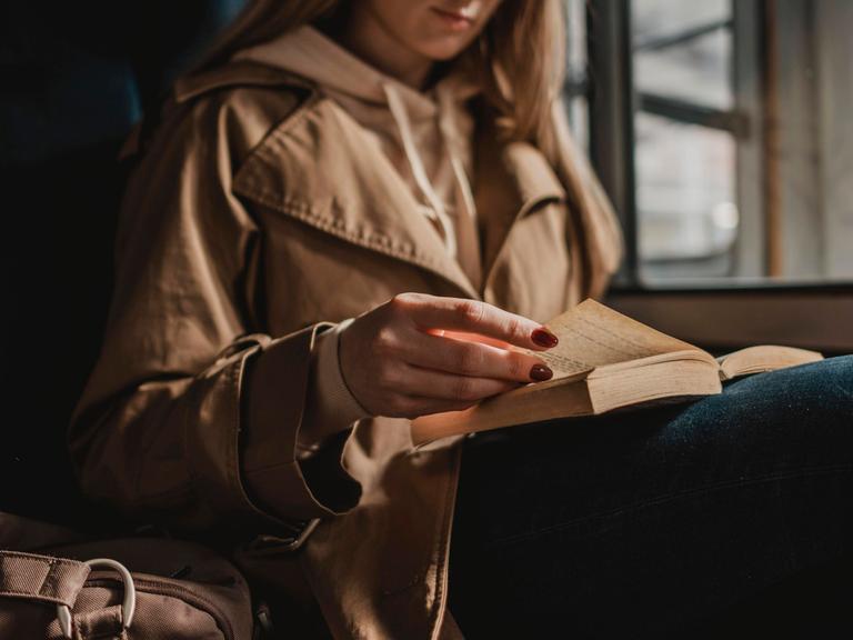 Frau mit hellbraunem Mantel sitzt in einem Zug und liest ein Buch im Zug Frau mit hellbraunem Mantel sitzt in einem Zug und liest ein Buch im Zug
