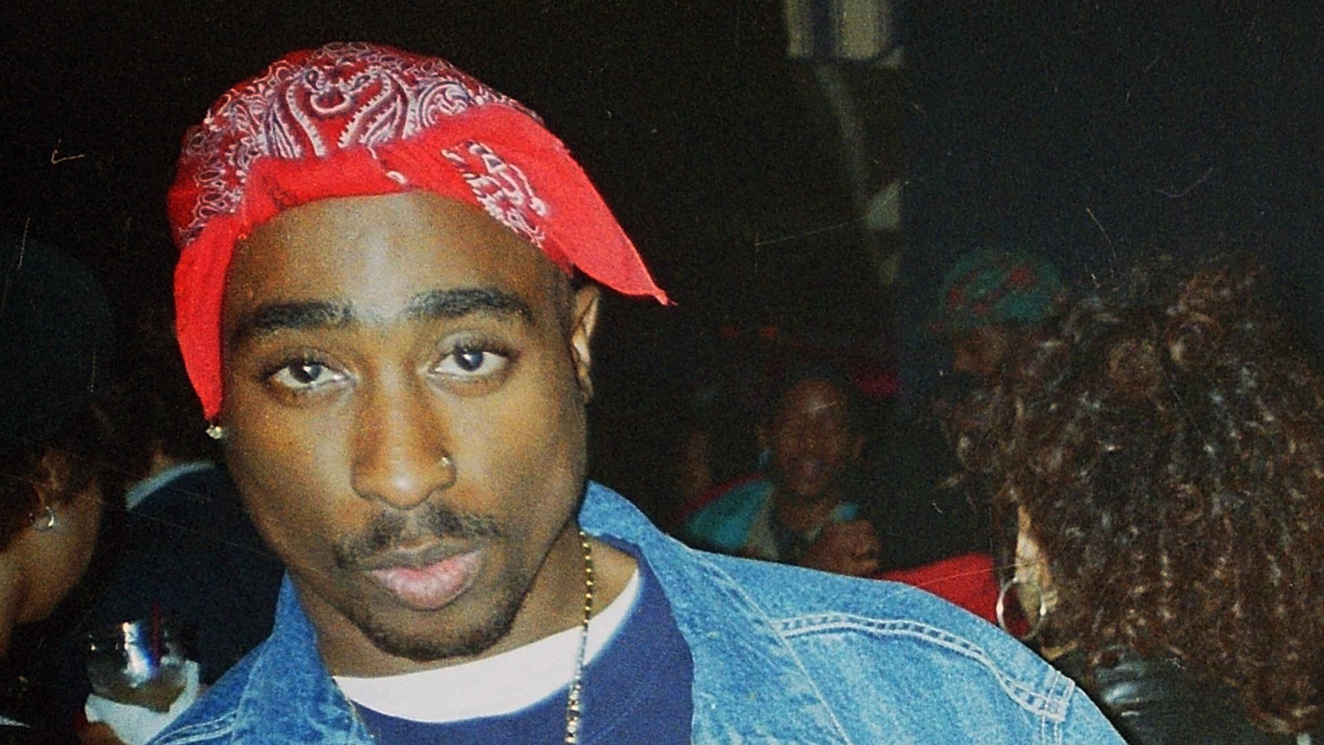 Ein junger Mann mit rotem Kopftuch schaut in die Kamera. Tupac Shakur trägt eine Jeansjacke , um den Hals hat er eine Halskette mit großem Kreuzanhänger. Die Mittelfinger streckt er an beiden Händen aus. Ein junger Mann mit rotem Kopftuch schaut in die Kamera. Tupac Shakur trägt eine Jeansjacke , um den Hals hat er eine Halskette mit großem Kreuzanhänger. Die Mittelfinger streckt er an beiden Händen aus.