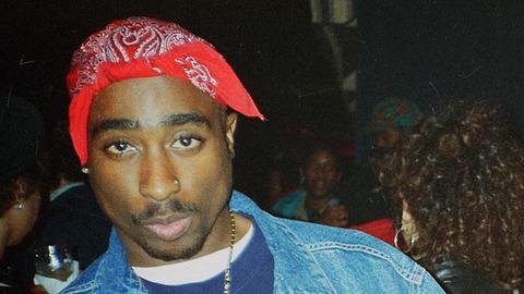 Ein junger Mann mit rotem Kopftuch schaut in die Kamera. Tupac Shakur trägt eine Jeansjacke , um den Hals hat er eine Halskette mit großem Kreuzanhänger. Die Mittelfinger streckt er an beiden Händen aus.