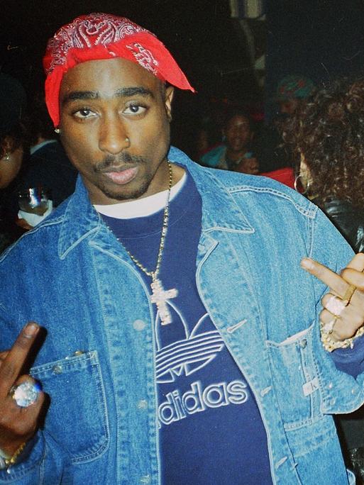 Ein junger Mann mit rotem Kopftuch schaut in die Kamera. Tupac Shakur trägt eine Jeansjacke , um den Hals hat er eine Halskette mit großem Kreuzanhänger. Die Mittelfinger streckt er an beiden Händen aus.