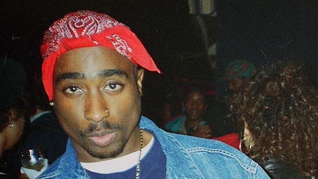 Ein junger Mann mit rotem Kopftuch schaut in die Kamera. Tupac Shakur trägt eine Jeansjacke , um den Hals hat er eine Halskette mit großem Kreuzanhänger. Die Mittelfinger streckt er an beiden Händen aus. Ein junger Mann mit rotem Kopftuch schaut in die Kamera. Tupac Shakur trägt eine Jeansjacke , um den Hals hat er eine Halskette mit großem Kreuzanhänger. Die Mittelfinger streckt er an beiden Händen aus.