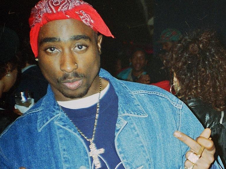 Ein junger Mann mit rotem Kopftuch schaut in die Kamera. Tupac Shakur trägt eine Jeansjacke , um den Hals hat er eine Halskette mit großem Kreuzanhänger. Die Mittelfinger streckt er an beiden Händen aus.