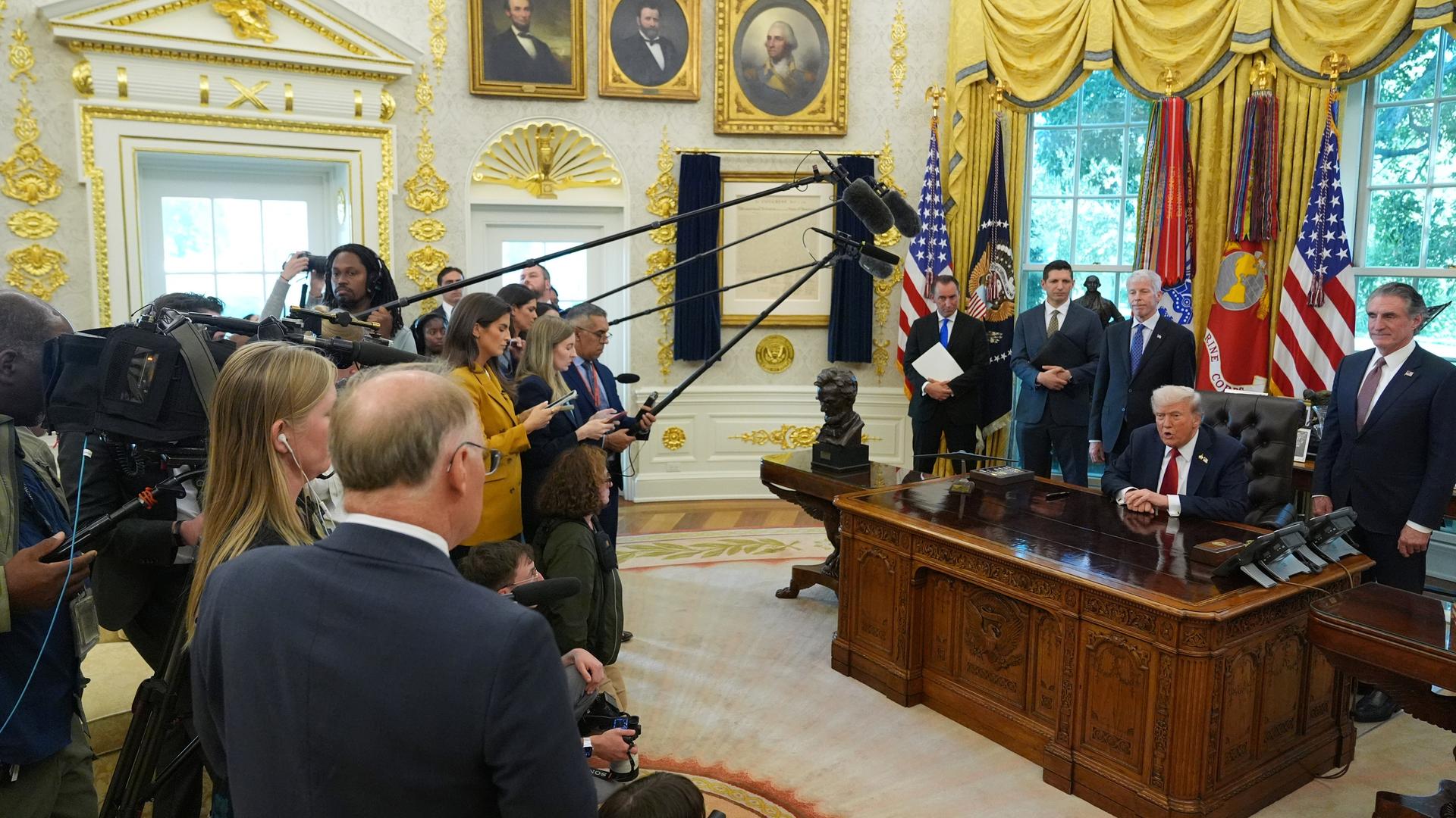 US-Präsident Trump spricht im Oval Office des Weißen Hauses mit Reportern. Trump sitzt an seinem Schreibtsich und mehrere Reporter halten Mikrofone in seine Richtung.