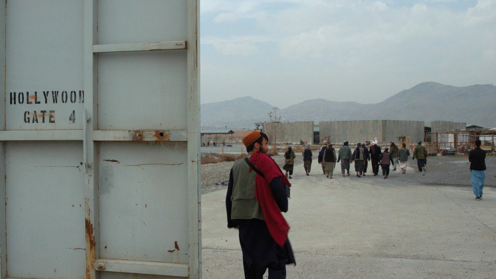 Der Eingang zu einem ehemaligen amerikanischen Lager. In der linken Bildhälfte die massive Lagertür, auf der in typischer Militärschrift das Wort "Hollywoodgate" steht, das Tor zu Hollywood. Rechts im Vordergrund ein Taliban, dahinter mehrere andere. Der Eingang zu einem ehemaligen amerikanischen Lager. In der linken Bildhälfte die massive Lagertür, auf der in typischer Militärschrift das Wort "Hollywoodgate" steht, das Tor zu Hollywood. Rechts im Vordergrund ein Taliban, dahinter mehrere andere.