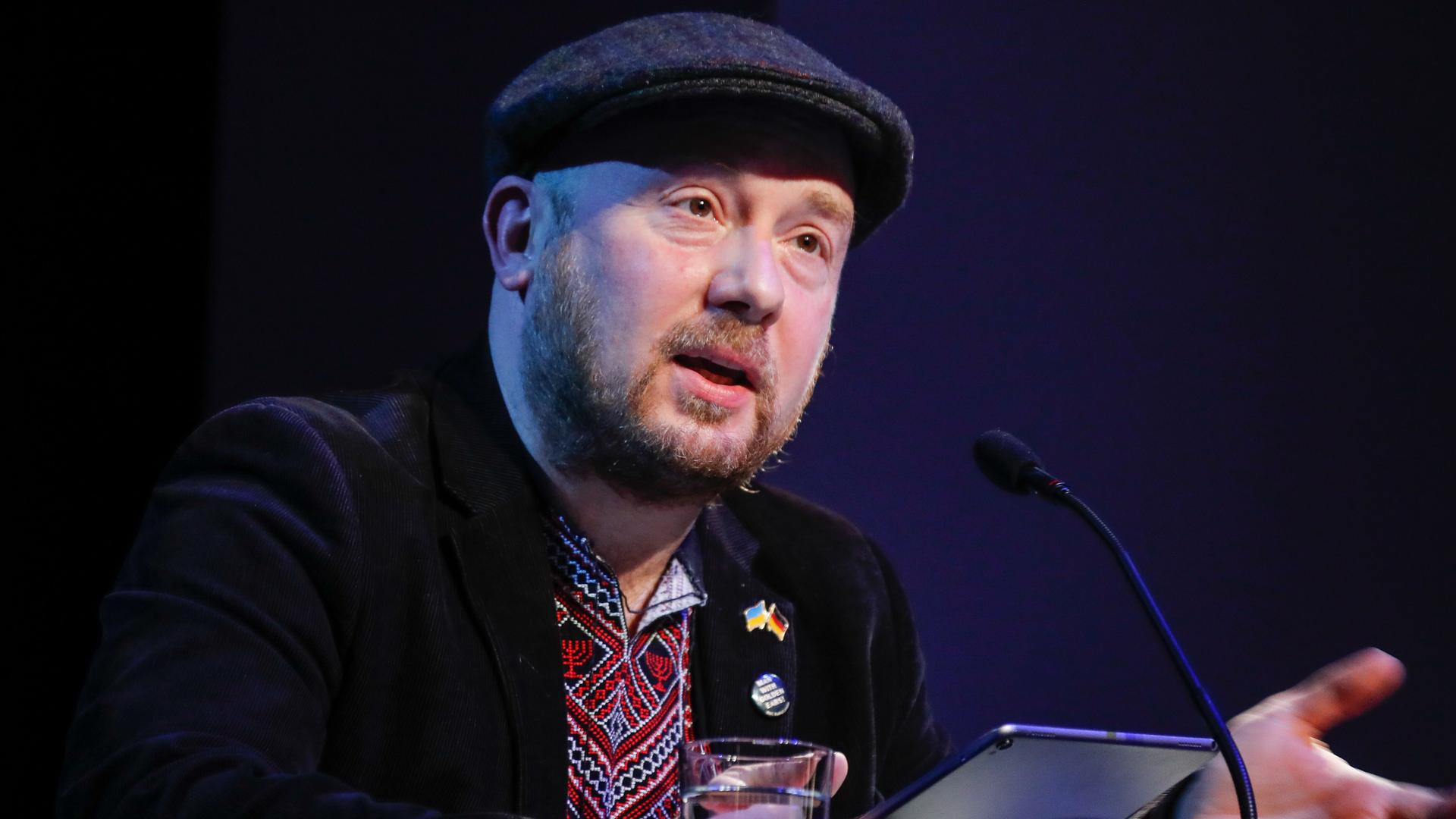 Yuriy Gurzhy sitzt mit Schiebermütze bei "Sprachlos die Sprache verteidigen: Lesung für die Ukraine" im Maxim Gorki Theater vor einem Mikrofon. Yuriy Gurzhy sitzt mit Schiebermütze bei "Sprachlos die Sprache verteidigen: Lesung für die Ukraine" im Maxim Gorki Theater vor einem Mikrofon.
