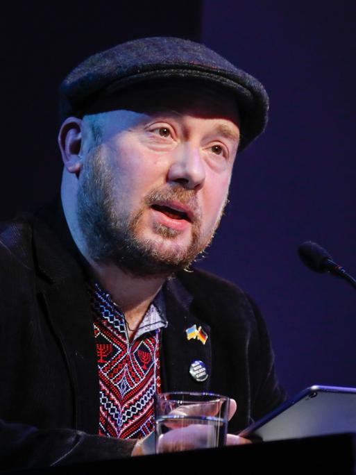Yuriy Gurzhy sitzt mit Schiebermütze bei "Sprachlos die Sprache verteidigen: Lesung für die Ukraine" im Maxim Gorki Theater vor einem Mikrofon.