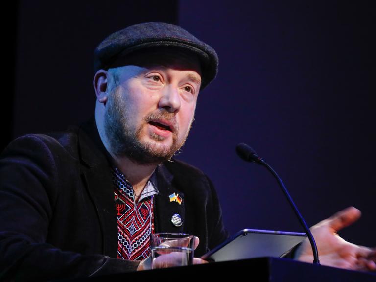 Yuriy Gurzhy sitzt mit Schiebermütze bei "Sprachlos die Sprache verteidigen: Lesung für die Ukraine" im Maxim Gorki Theater vor einem Mikrofon.