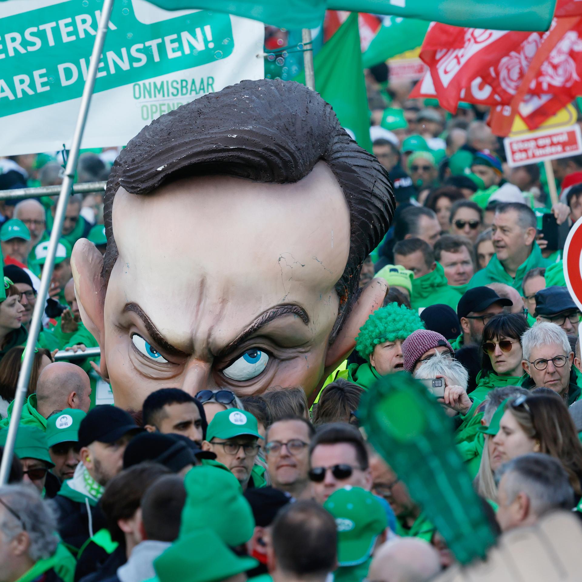 Viele Menschen, die in grün gekleidet sind, halten Schilder hoch. In der Mitte sieht man einen Puppenkopf, der Regierungschef Bart de Wever darstellen soll.
