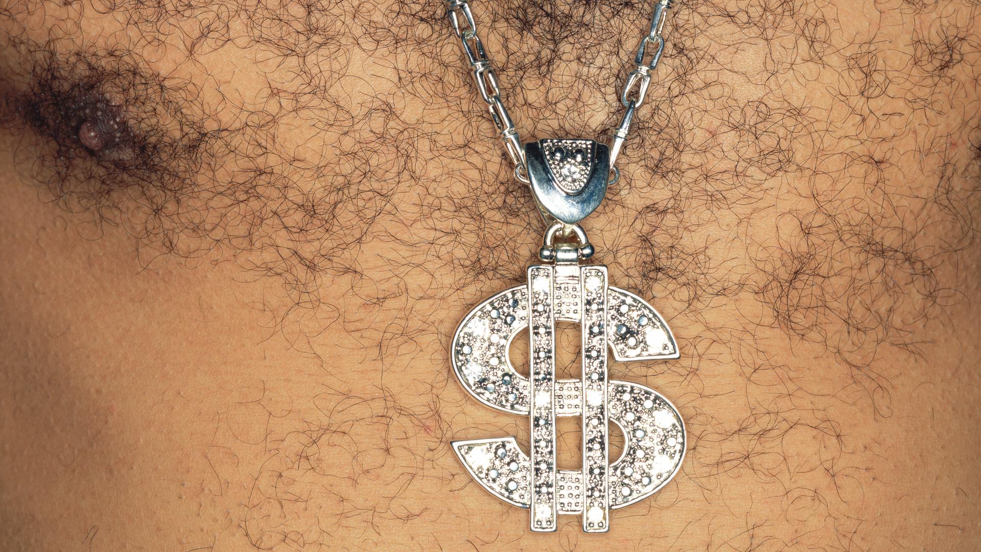 Ein Mann mit behaarter Brust, der ein glitzernde Kette mit einem US-Dollar-Zeichen als Anhänger trägt. Ein Mann mit behaarter Brust, der ein glitzernde Kette mit einem US-Dollar-Zeichen als Anhänger trägt.