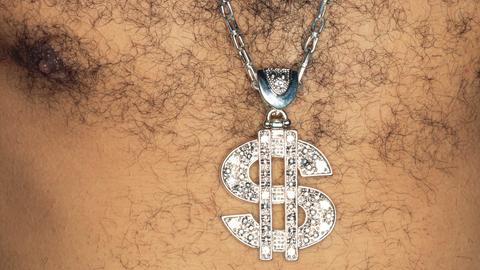 Ein Mann mit behaarter Brust, der ein glitzernde Kette mit einem US-Dollar-Zeichen als Anhänger trägt.