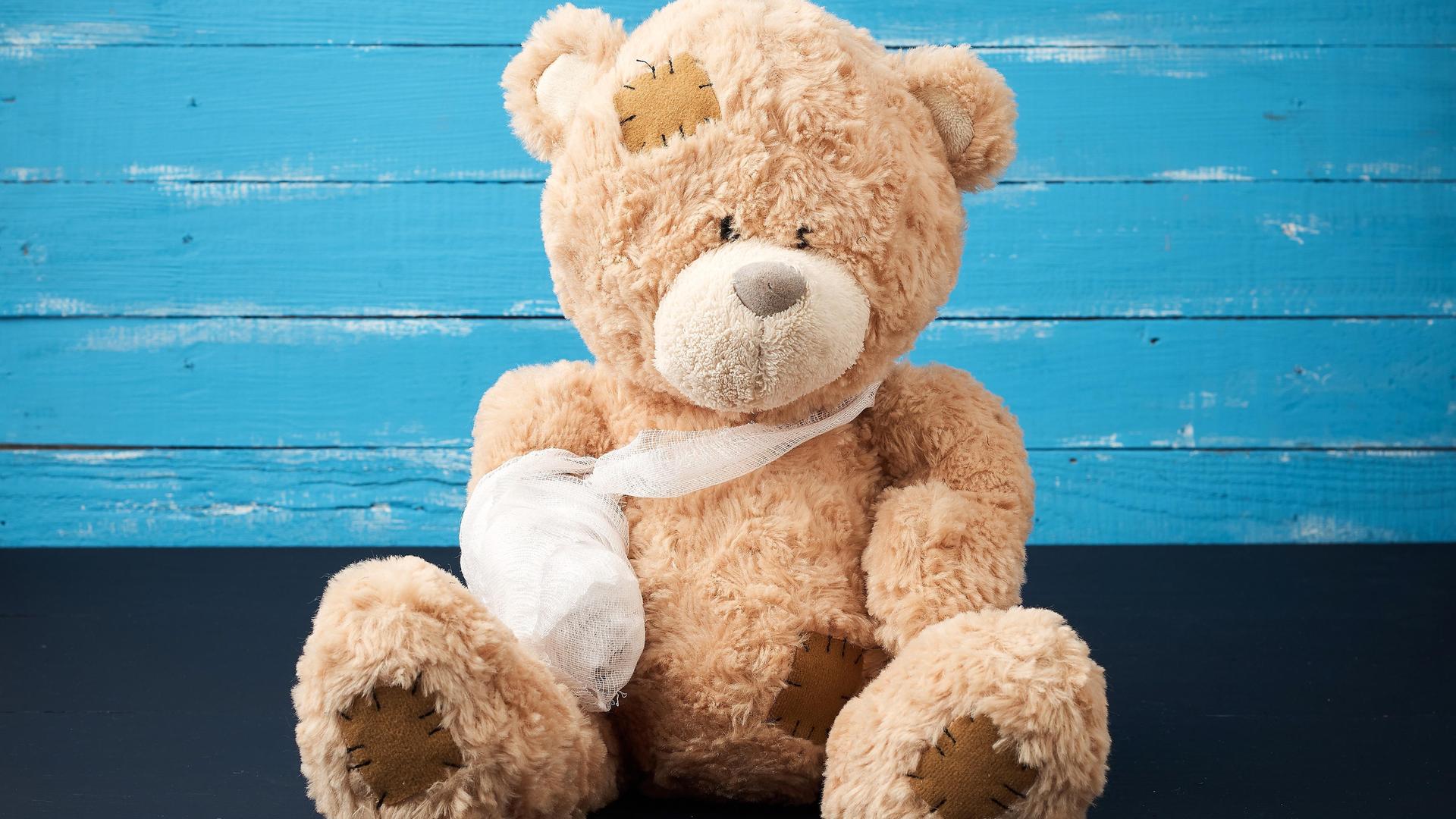 Ein brauner Teddy sitzt vor einer blauen Wand, seiner einer Arm hängt in einer Schlinge und ist eingegipst.