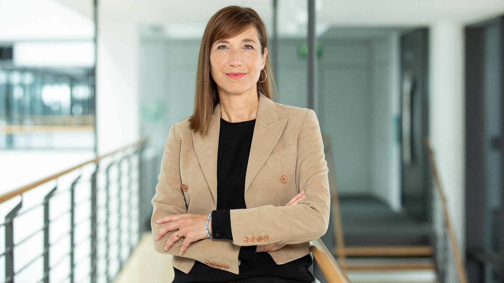 Barbara Massing steht in einem offenen Treppenhaus, sie hält die Arme verschränkt.
