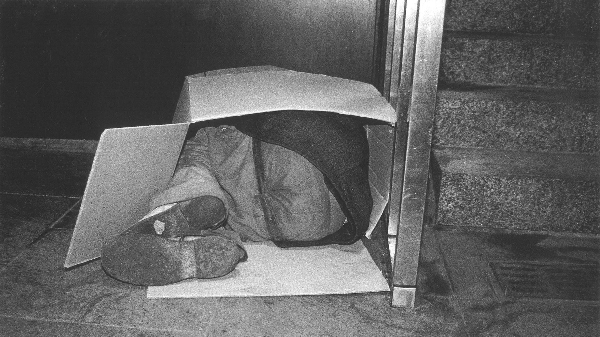 Schwarzweißfoto einer Person, die in Embryonalstellung in einem Karton liegt.