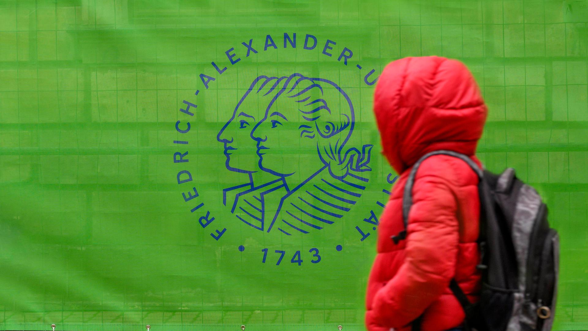 Eine Person im roten Anorak geht an einem Bauzaun vorbei, der mit einem grünen Transparent mit der Aufschrift "Friedrich Alexander Universität 1743" verkleidet ist.