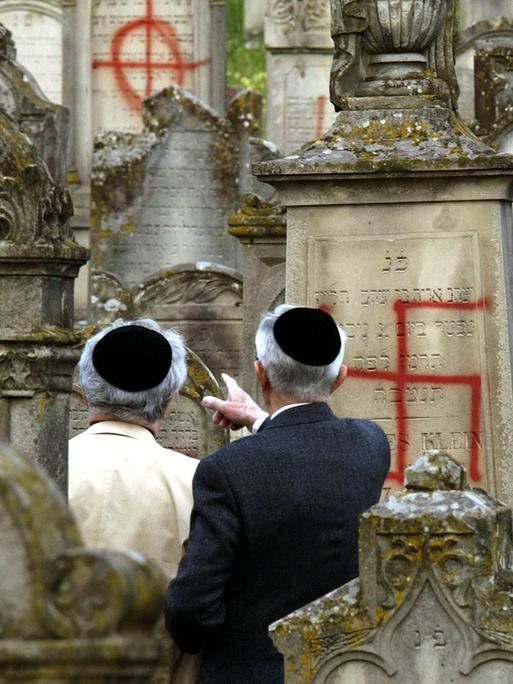 Zwei Männer mit Kippa auf einem jüdischen Friedhof. Einige Grabsteine sind mit Hakenkreuzen beschmiert worden.