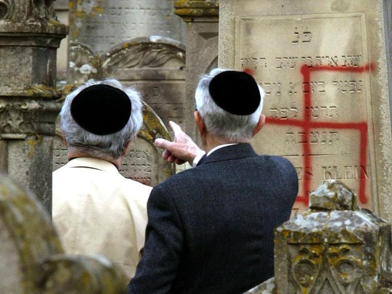 Zwei Männer mit Kippa auf einem jüdischen Friedhof. Einige Grabsteine sind mit Hakenkreuzen beschmiert worden.