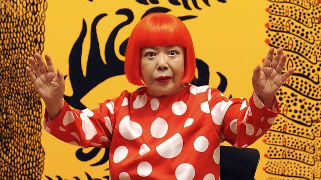 Zu sehen ist die japanische Künstlerin Yayoi Kusama. Sie trägt eine rote Perücke und ein Kleid mit weißen Punkten. Das Foto entstand im Jahr 2012 vor einer Ausstellung in der Stadt London im Land Großbritannien.