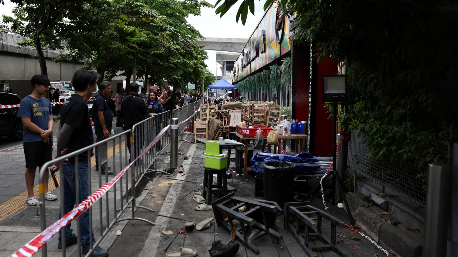 Personen stehen hinter einer Absperrung des Brandortes der Bar „Rong Beer Na Lat Phrao“ in Bangkok. Mobiliar liegt auf dem Boden. Personen stehen hinter einer Absperrung des Brandortes der Bar „Rong Beer Na Lat Phrao“ in Bangkok. Mobiliar liegt auf dem Boden.