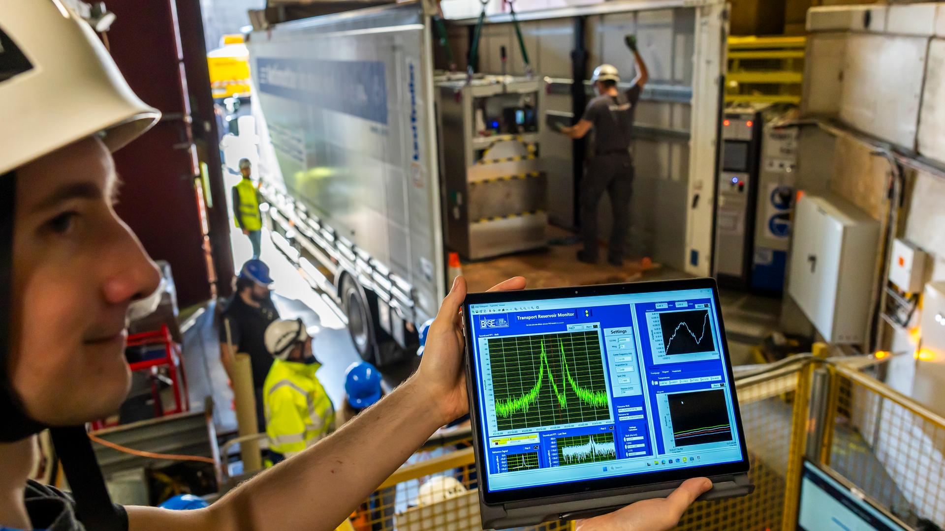 Ein Wissenschaftler am CERN zeigt einen Bildschirm, auf dem Antimateriemenge zu sehen ist in Form von Kurven. Im Hintergrund wird eine Box in einen LKW eingeladen. Europäische Organisation für Kernforschung in Genf im März 2026