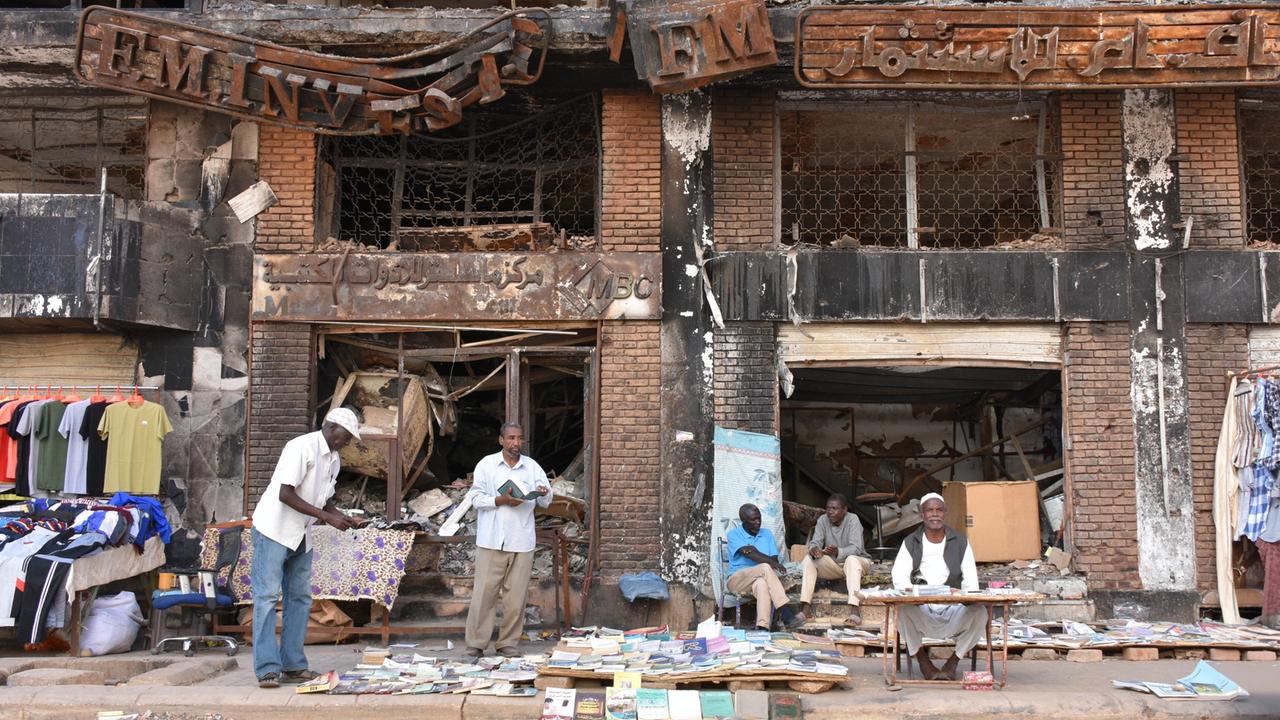 Einige Männer sitzen und stehen vor einem kriegsversehrten Haus in Sudans Hauptstadt Khartum und verkaufen Kleidung und Bücher. 