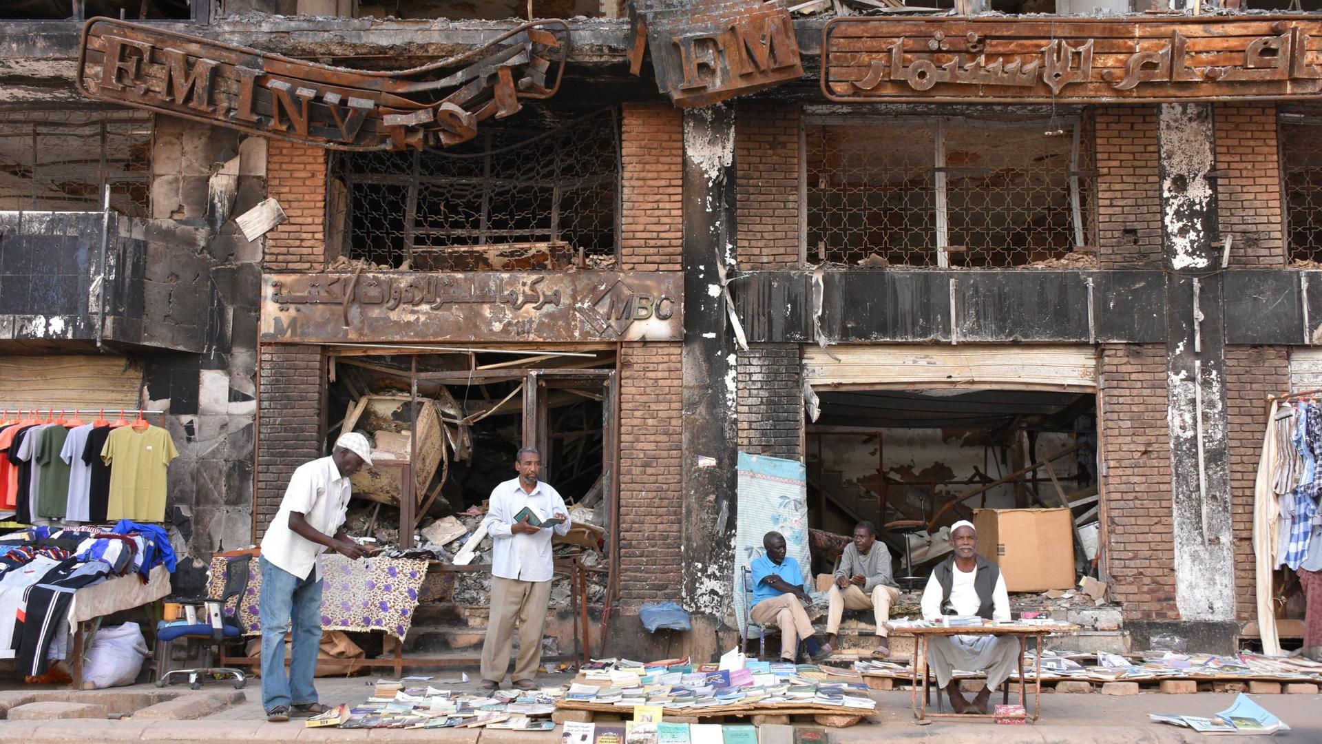 Einige Männer sitzen und stehen vor einem kriegsversehrten Haus in Sudans Hauptstadt Khartum und verkaufen Kleidung und Bücher. 