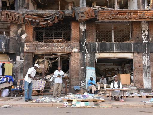 Einige Männer sitzen und stehen vor einem kriegsversehrten Haus in Sudans Hauptstadt Khartum und verkaufen Kleidung und Bücher. 