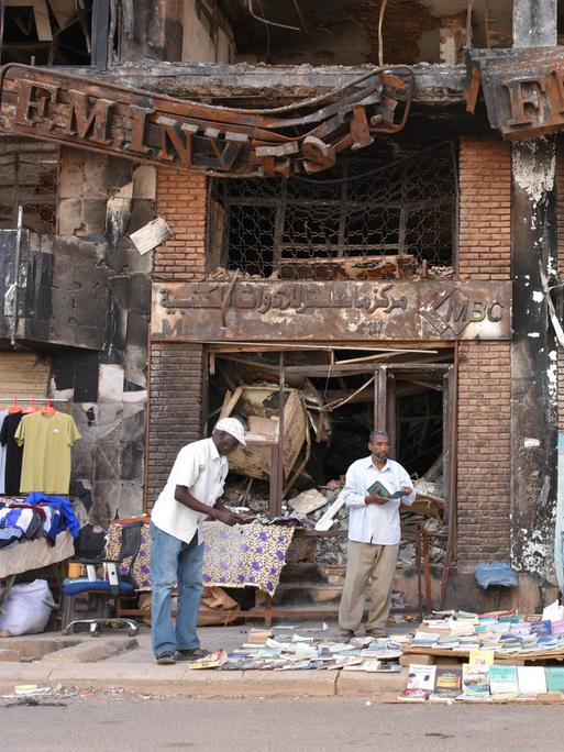 Einige Männer sitzen und stehen vor einem kriegsversehrten Haus in Sudans Hauptstadt Khartum und verkaufen Kleidung und Bücher. 
