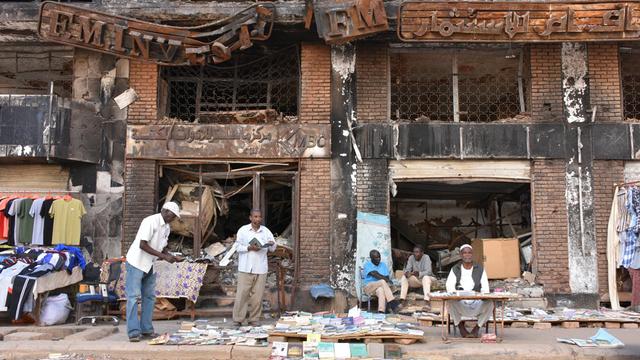 Einige Männer sitzen und stehen vor einem kriegsversehrten Haus in Sudans Hauptstadt Khartum und verkaufen Kleidung und Bücher. 