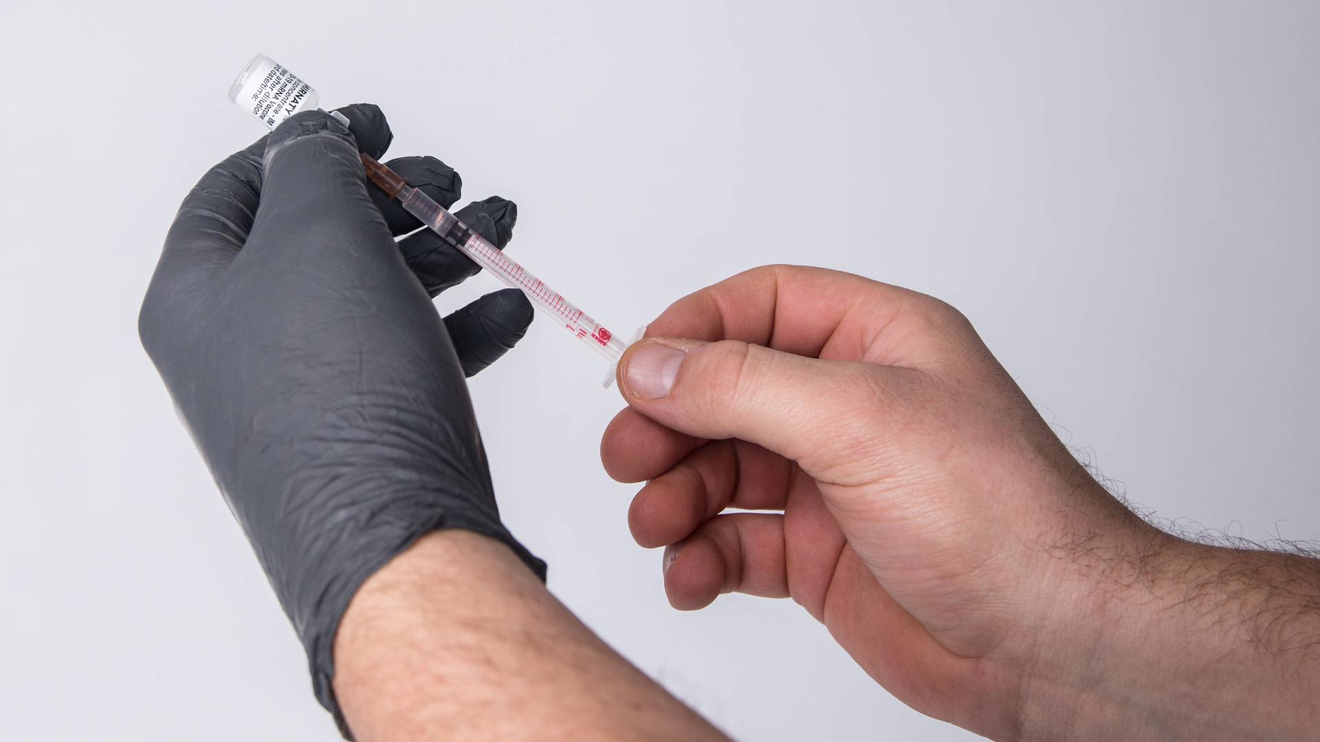Eine Person mit einem schwarzen Schutzhandschuh zieht mit einer Impfspritze eine Impfdosis auf.