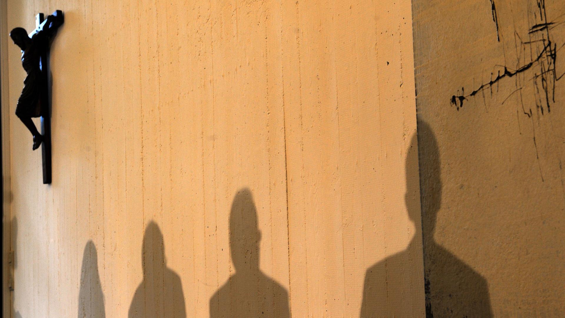 Schatten von Menschen auf einer kahlen Wand mit einem Kruzifix. 