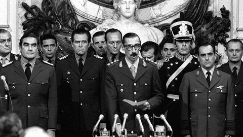 Ein Schwarz-Weiß-Foto zeigt General Jorge Rafael Videla, der in ein Mikrofon spricht. Er ist von mehreren hochrangigen argentinischen Militärangehörigen eingerahmt.