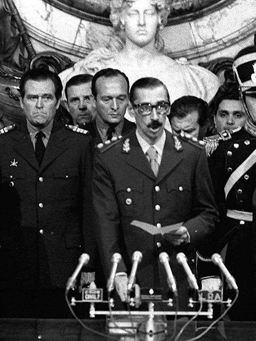 Ein Schwarz-Weiß-Foto zeigt General Jorge Rafael Videla, der in ein Mikrofon spricht. Er ist von mehreren hochrangigen argentinischen Militärangehörigen eingerahmt. Ein Schwarz-Weiß-Foto zeigt General Jorge Rafael Videla, der in ein Mikrofon spricht. Er ist von mehreren hochrangigen argentinischen Militärangehörigen eingerahmt.