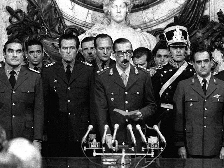 Ein Schwarz-Weiß-Foto zeigt General Jorge Rafael Videla, der in ein Mikrofon spricht. Er ist von mehreren hochrangigen argentinischen Militärangehörigen eingerahmt. Ein Schwarz-Weiß-Foto zeigt General Jorge Rafael Videla, der in ein Mikrofon spricht. Er ist von mehreren hochrangigen argentinischen Militärangehörigen eingerahmt.