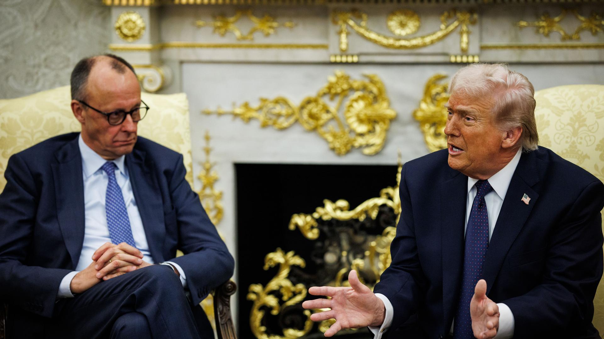 Bundeskanzler Friedrich Merz sitzt neben US-Präsident Donald Trump, der gerade spricht und gestikuliert, in goldenen Sesseln vor einem goldbeschlagenen Kamin im Oval Office im Weißen Haus in Washington.  