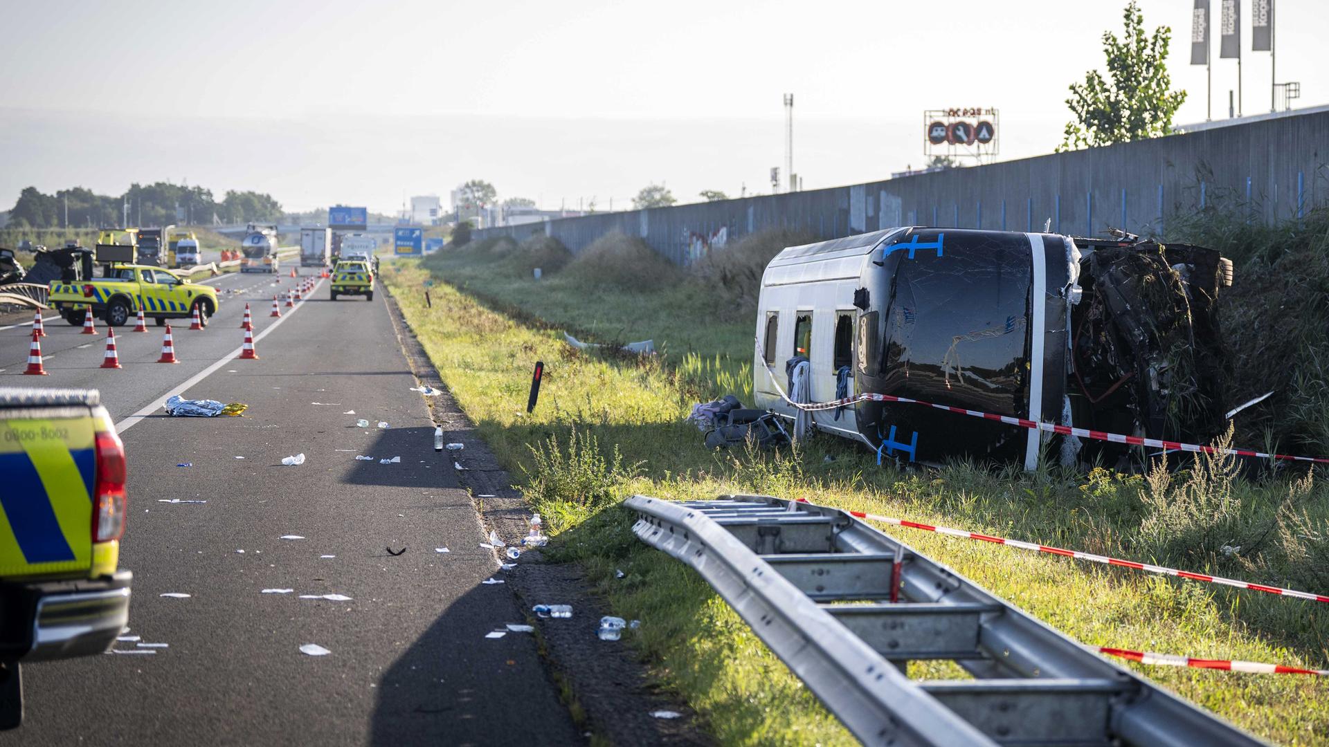 Der Bus liegt auf der Seite gekippt an einer Lärmschutzwand am Rande der Autobahn, die durch Warnkegel abgesperrt ist. Auf der Fahrbahn liegen Wrackteile. Mehrere Polizeifahrzeuge stehen auf der Autobahn. Der Bus liegt auf der Seite gekippt an einer Lärmschutzwand am Rande der Autobahn, die durch Warnkegel abgesperrt ist. Auf der Fahrbahn liegen Wrackteile. Mehrere Polizeifahrzeuge stehen auf der Autobahn.
