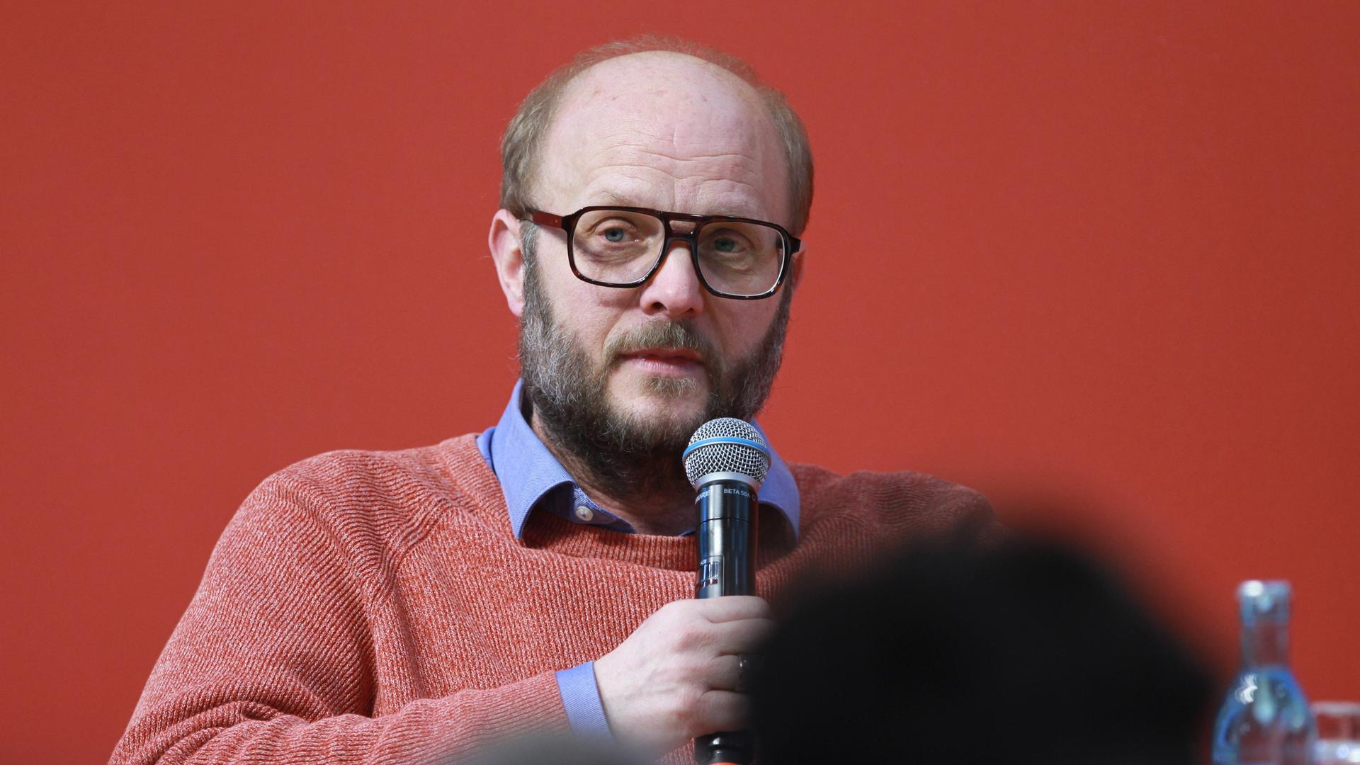 David Schraven sitzt vor einer roten Wand, hält ein Mikrofon in der Hand und scheint mit dem Publikum zu sprechen