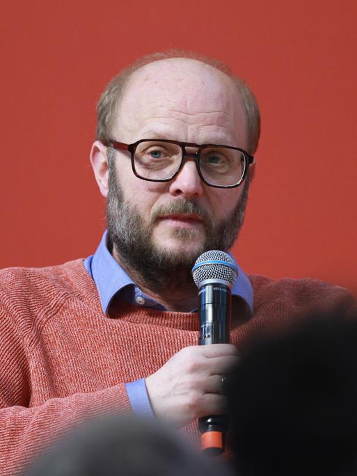 David Schraven sitzt vor einer roten Wand, hält ein Mikrofon in der Hand und scheint mit dem Publikum zu sprechen